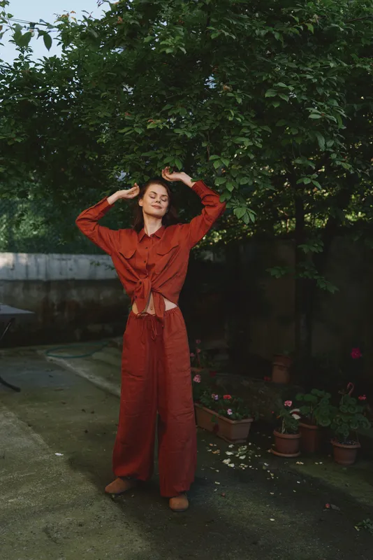 Cheerful woman outdoors in a red outfit poses with relaxed stance in a sunlit garden, natural tones, soft breeze, candid lifestyle moment