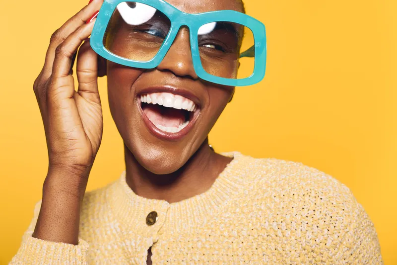 Happy dark skinned African woman wearing oversized blue glasses laughs joyfully on yellow background. Close-up portrait with natural makeup and casual knitted sweater, concept of fun and fashion.