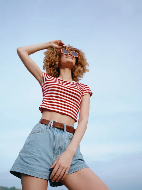 Young woman in a red and white striped crop top and high waisted denim shorts stands confidently under a clear blue sky, exuding carefree summer style.