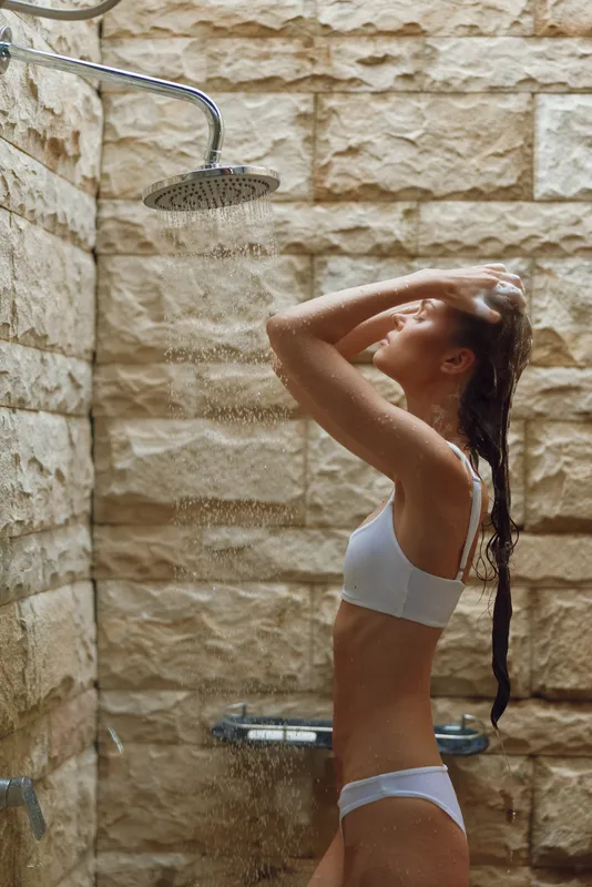 Young woman in white swimwear enjoying an outdoor shower with water streaming down, natural stone wall background, serene and refreshing moment captured.