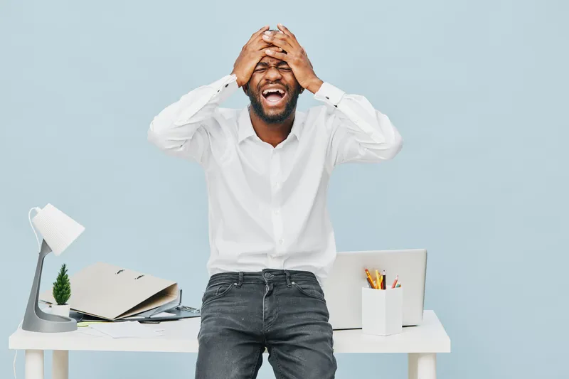 Joyful man with beard in white shirt stands behind office desk, covering face with hands, expressing frustration or stress, in a bright modern workspace with pastel blue background. People lifestyle c