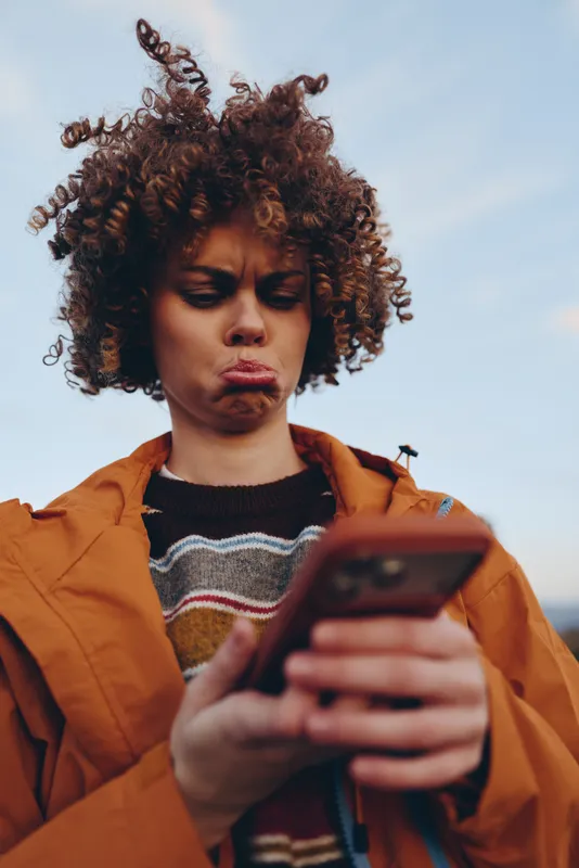 Woman lifestyle outdoors, curly hair, orange jacket, holding smartphone, focused gaze, golden daylight, technology moment, relaxed posture, soft breeze, sky background, communication
