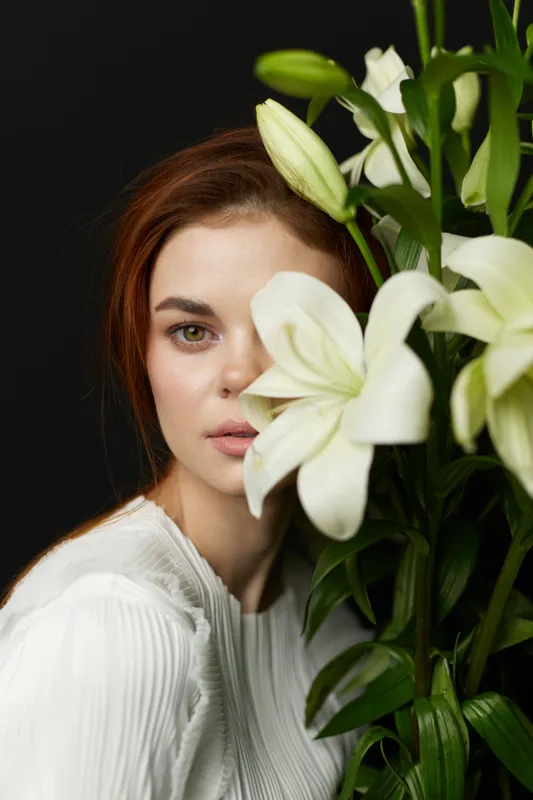 Beautiful young woman with long hair and natural makeup, partially hidden by white lilies, exuding calmness and elegance against a dark background