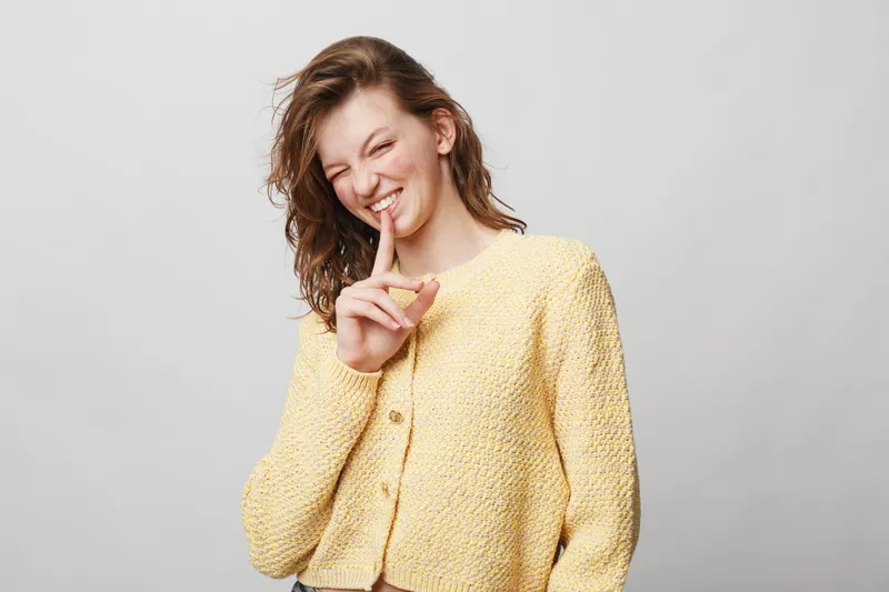 Young woman in yellow knitted sweater smiles and holds finger to lips in a shushing gesture. Studio portrait on light background with playful and secretive mood.