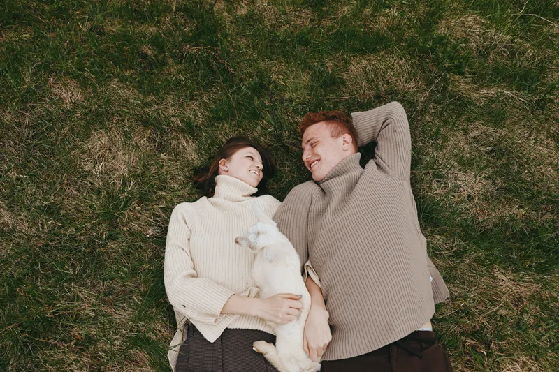 Cheerful couple outdoors, lying on grass with a playful goat kid between them, warm light, cozy sweaters, relaxed smiles, natural colors, tranquil moment, gentle breeze, connection, carefree mood