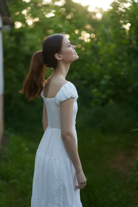 Young woman in a white dress enjoying nature, smiling in a serene green environment, capturing the essence of joy and tranquility in summer