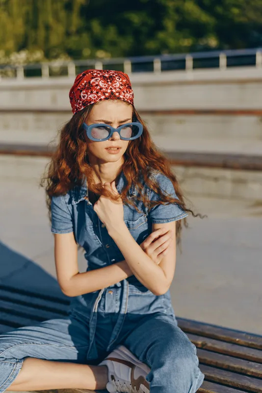 Lifestyle woman in modern boho-western denim street style wearing blue sunglasses and red bandana, captured with warm film color tones outdoors on a sunny day.