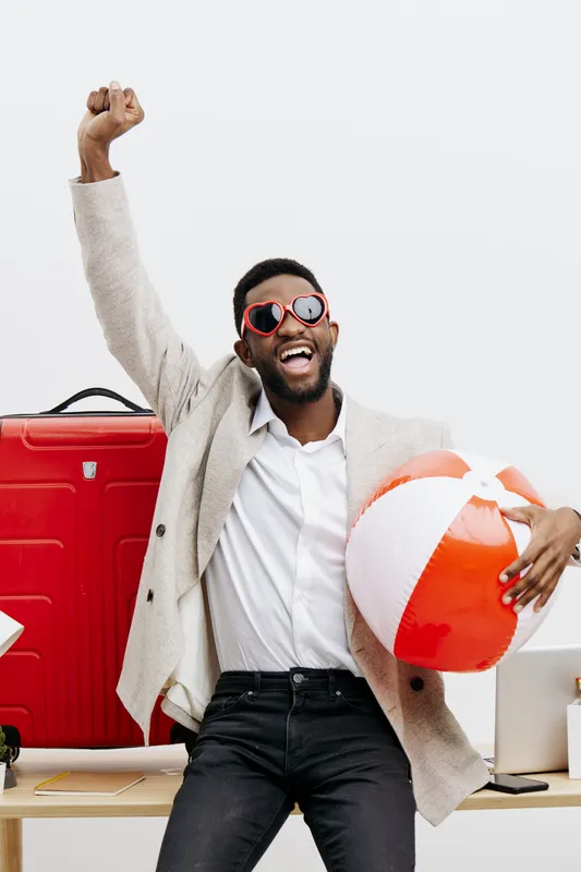 Cheerful stylish man of African descent with beard and glasses celebrating with summer vacation items including a beach ball and suitcase, in casual clothing, indoors on white background. People lifes