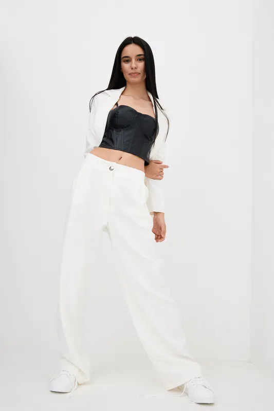 young woman in white pants and black corset stands confidently posing in bright studio with minimalistic white background and casual streetwear style