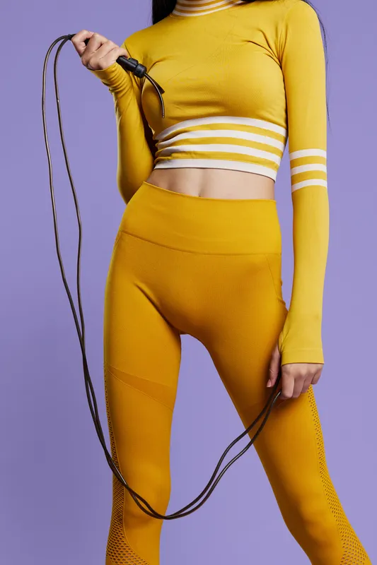 Athletic woman sport fitness holding jump rope, dressed in stylish yellow activewear, showcasing strength, agility, and wellness on a soft purple background.