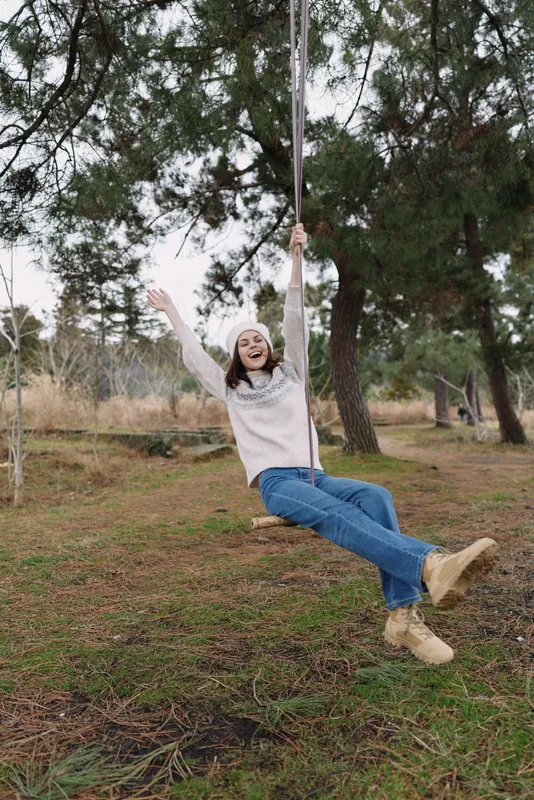 Woman on a swing in an outdoor park, joyfully swinging among trees, wearing a sweater and boots for a casual leisure mood.