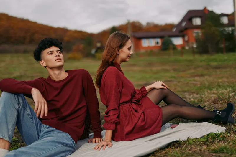 Candid portrait of a young couple seated back-to-back on a blanket in an autumn meadow, conveying authenticity and credibility through natural poses, warm light and sincere expressions.