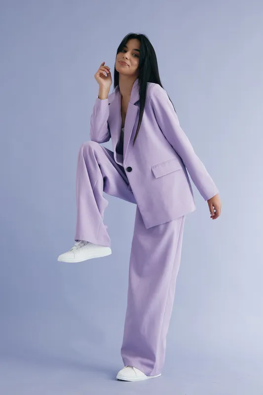 Woman in stylish lilac suit posing confidently against a soft purple backdrop, wearing white sneakers, showcasing modern fashion and relaxed attitude.