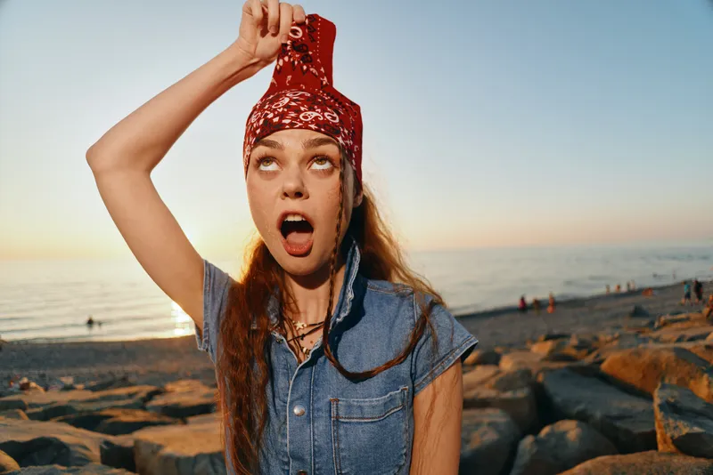 Young woman in modern boho-western denim street style holds red bandana over head with playful expression, set against beach at sunset, film color effect enhancing mood.