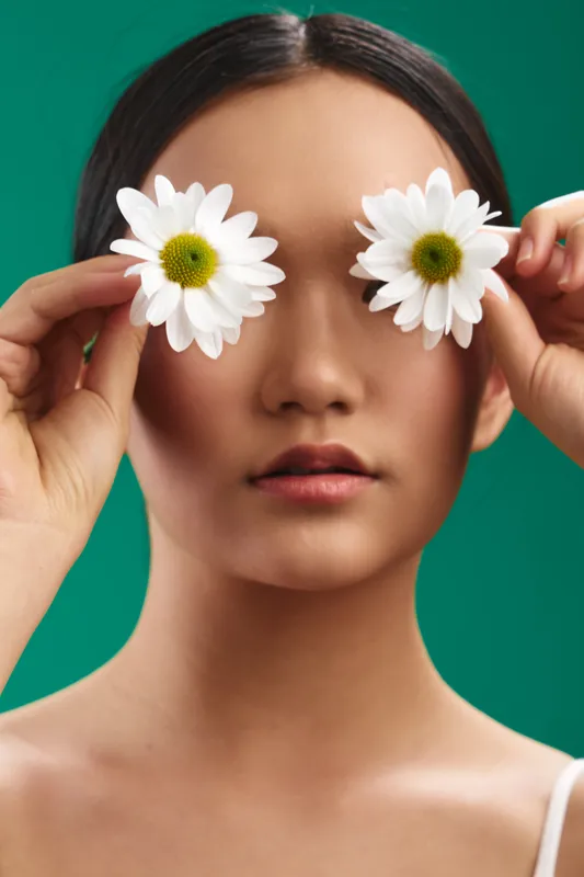 asian woman holds flowers over her eyes, creating a playful portrait in a vibrant studio. the image conveys whimsy, charm, and creative beauty with a bright mood.