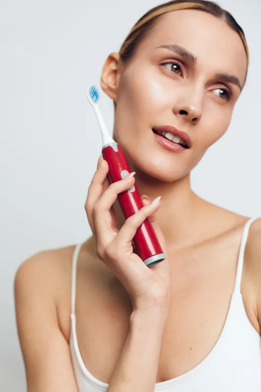 Young woman holds electric toothbrush close to face with thoughtful expression wearing white tank top isolated on light background, personal care and oral hygiene concept.