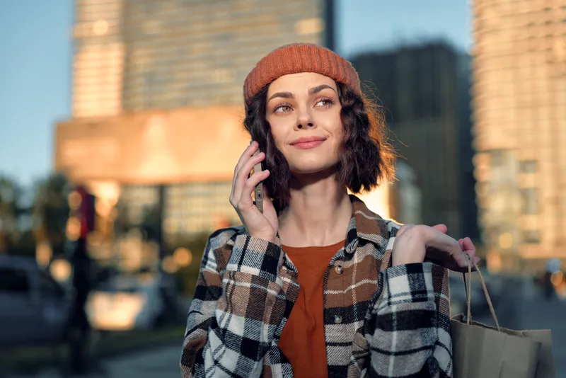 Woman urban shopping portrait with phone and beanie, smiling in golden hour glow on city street. Authenticity, candid lifestyle, mindful living and emotional storytelling