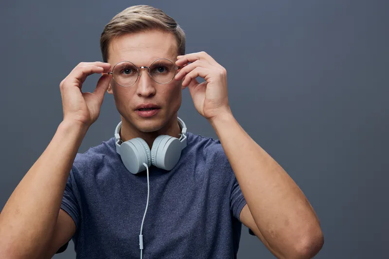 Close-up of a young man with glasses adjusting them, wearing headphones around his neck, looking serious and focused, against a plain gray background. Studio portrait of a male with short hair, casual