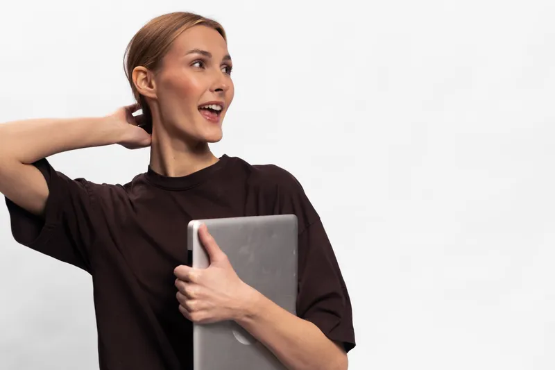 Young woman in casual dark shirt holding laptop looks aside with smile on white background. Concept for technology, remote work, education, and happy lifestyle.