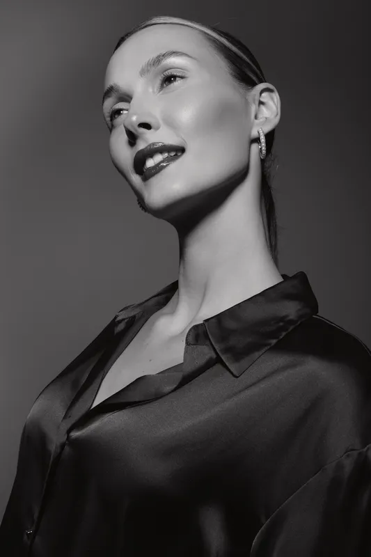 Elegant white woman with light skin and slicked back hair poses in shiny blouse, looking upward with confident smile. Black and white portrait with studio lighting and minimalistic style.