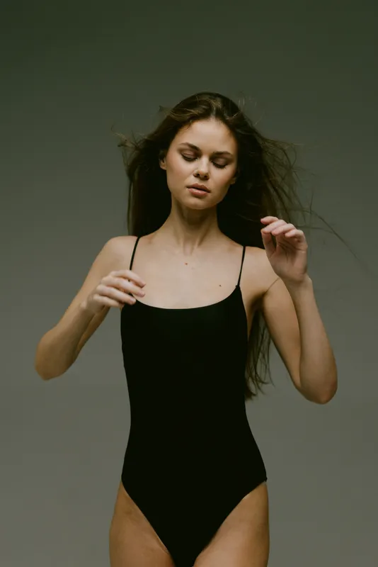 Young woman in black swimsuit with closed eyes posing against gray background, calm expression, minimalistic style