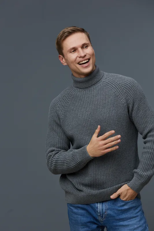 Smiling young man wearing cozy gray sweater and jeans, looking to the side and laughing against plain dark background, studio portrait. People lifestyle concept, casual fashion, happiness.