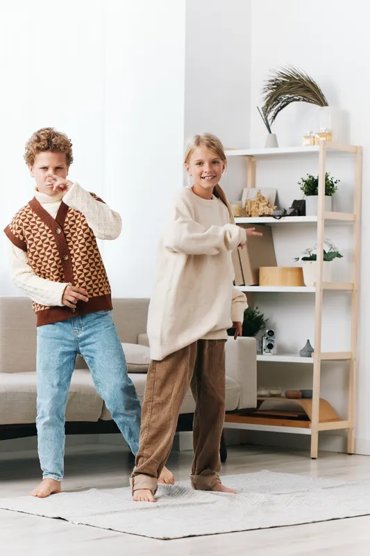 Two children dance playfully indoors wearing cozy knitwear and loungewear, capturing candid joy and movement in a bright, minimal living space and warm home atmosphere.