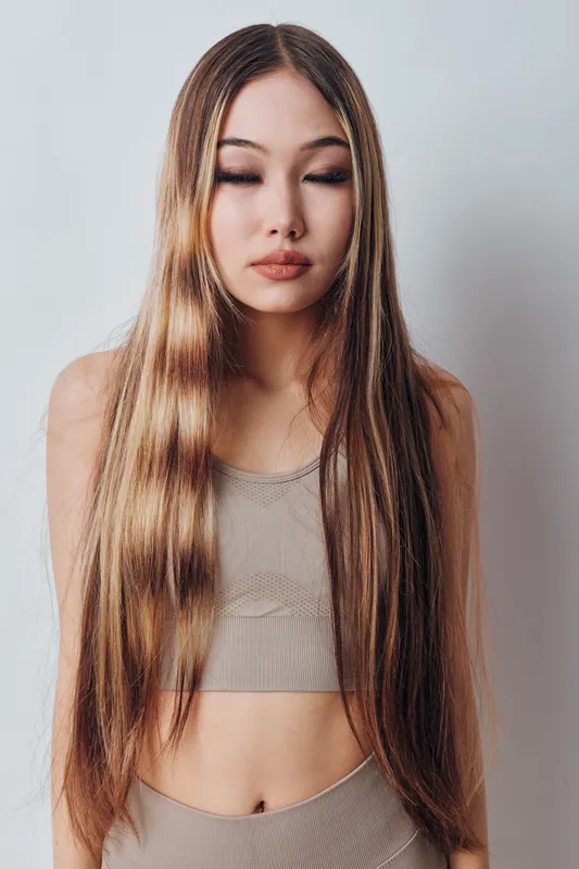 Young woman with long straight hair and closed eyes wearing neutral tone sleeveless top standing against white background. Beauty, fashion and calm expression with natural style.