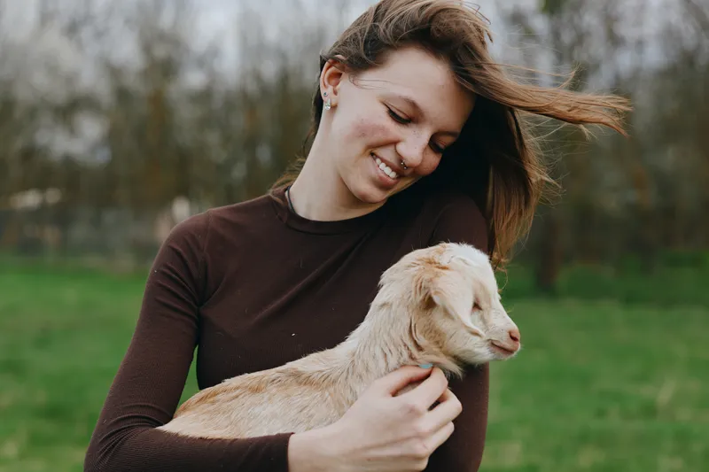 A woman warmly embraces a small goat kid in a green field, smiling with affection, enjoying a simple outdoor lifestyle, gentle moment between human and animal, peaceful rural scene.