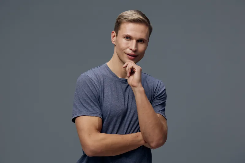 Confident young man with light brown hair wearing casual blue T-shirt looking at camera posing against gray background studio portrait. People lifestyle concept