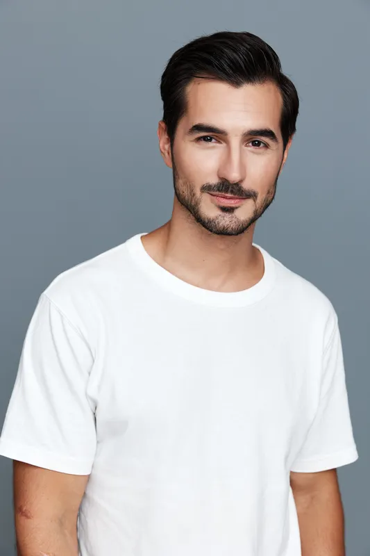 Close-up portrait of a smiling man with dark hair wearing a plain white t-shirt against a solid gray background. Confidence and positivity radiate from his expression.