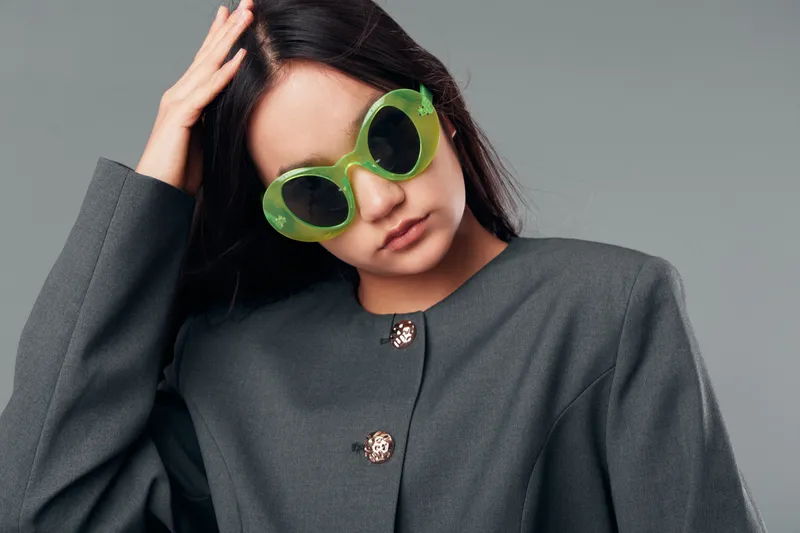 Young woman with long dark hair wears oversized green sunglasses and grey blazer posing with hand on head against grey background in studio. Fashion, style and portrait concept.
