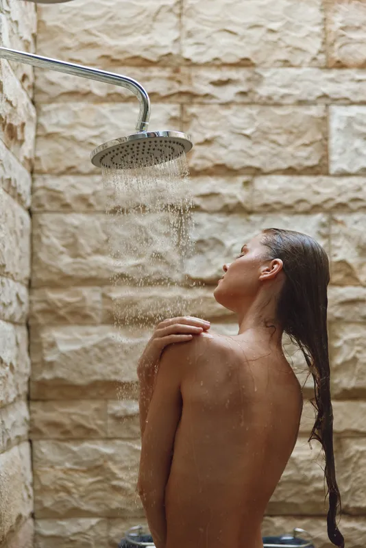 woman showering outdoors under rainfall showerhead with natural stone wall, relaxed pose, wet hair flowing, serene and refreshing moment in nature