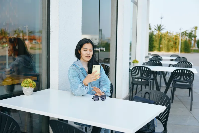 Woman at an outdoor cafe uses smartphone, white table, sunglasses beside, casual attire, relaxed urban dining scene, daytime leisure, connected moment, customer creates digital interaction