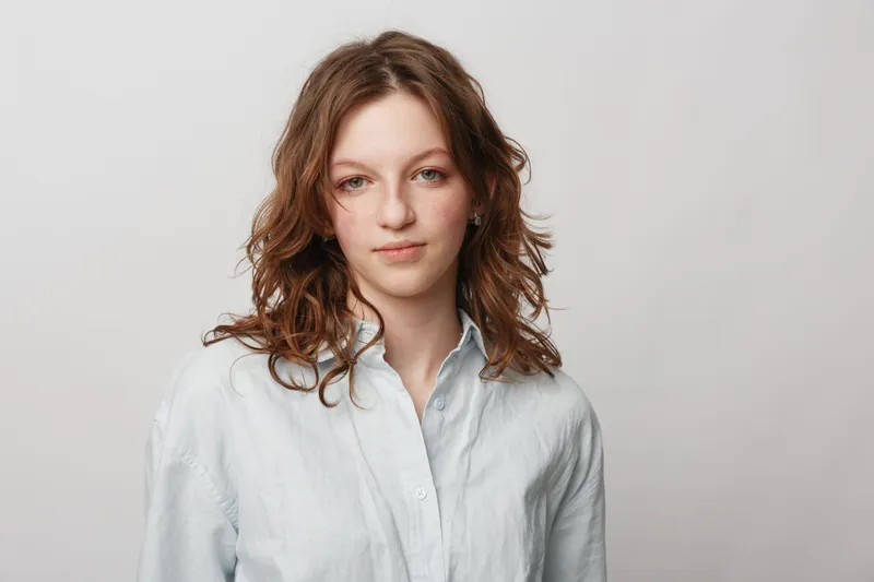 Young woman with curly brown hair wearing a white shirt looks confidently at camera on light background. Portrait of casual style and natural beauty concept.