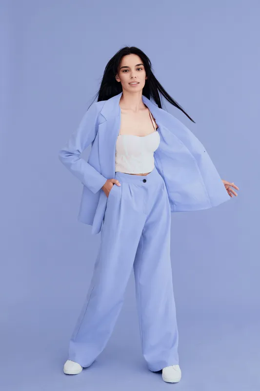 Woman wearing pastel blue suit stands confidently against light purple background, showcasing modern fashion style with relaxed pose and natural expression.