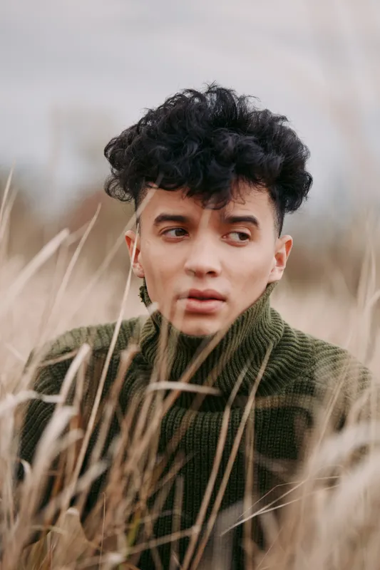 A young person in a textured green sweater peers through tall dried grasses in a candid close-up portrait, conveying authentic emotion and natural expression with muted earthy tones.