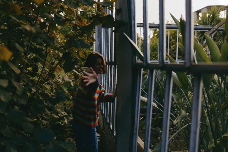 Child girl in a colorful striped sweater reaching hand through a metal gate, curious and smiling, about seven years old, leaning among tropical plants and dense green foliage at dusk.