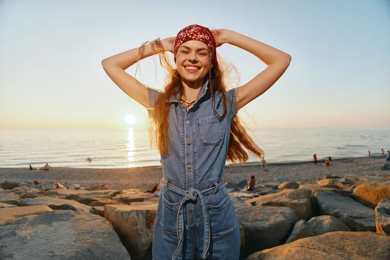smiling woman in denim jumpsuit with red bandana enjoying sunset on rocky beach with ocean background and golden light