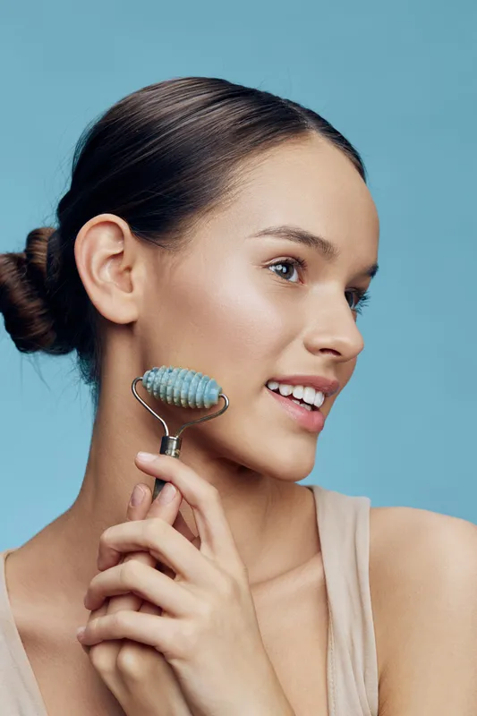 Young woman with a jade roller smiling against a blue background, promoting skincare, self care, and beauty routines with a fresh and vibrant look