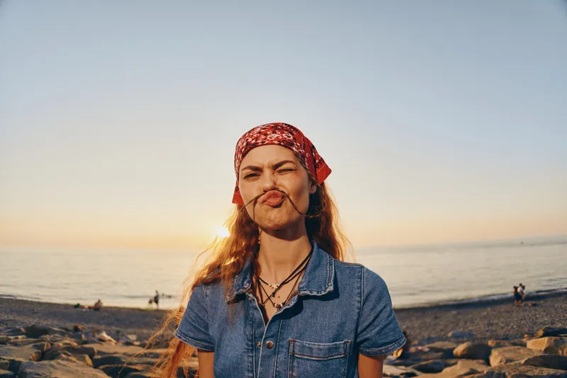 Lifestyle woman in modern boho-western denim street style with film color effect making a playful face outdoors near the beach during golden hour.
