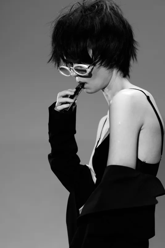 Black and white portrait of a stylish woman with short hair wearing sunglasses and a black outfit, expressing thoughtful mood in minimalistic setting.