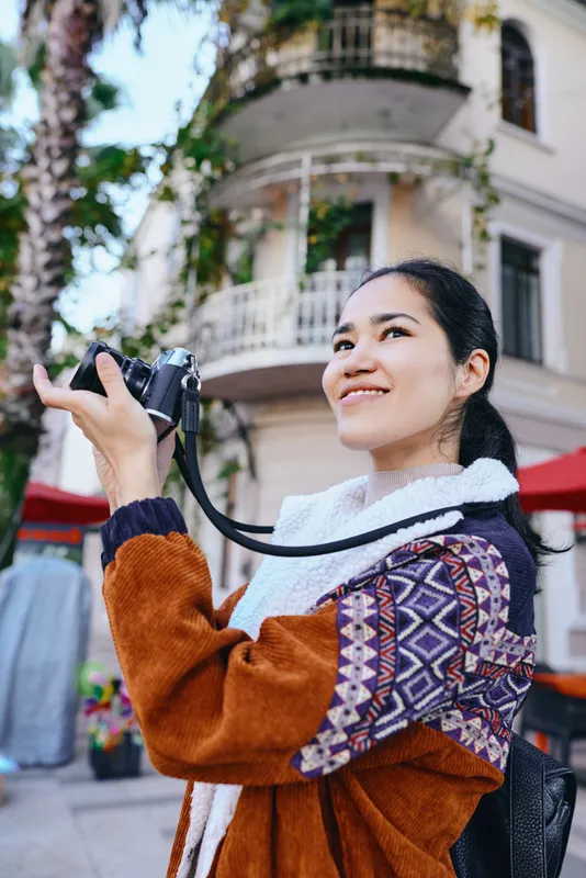 Woman with camera exploring urban street in sunny outdoor setting, fashion jacket and patterned sleeves, capturing candid moments and city textures for editorial travel and lifestyle storytelling,