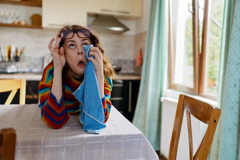 Casual woman indoors, in a cozy kitchen, wiping her eye with a towel, wearing a colorful striped sweater and glasses on her head, celebrating a relaxed home lifestyle