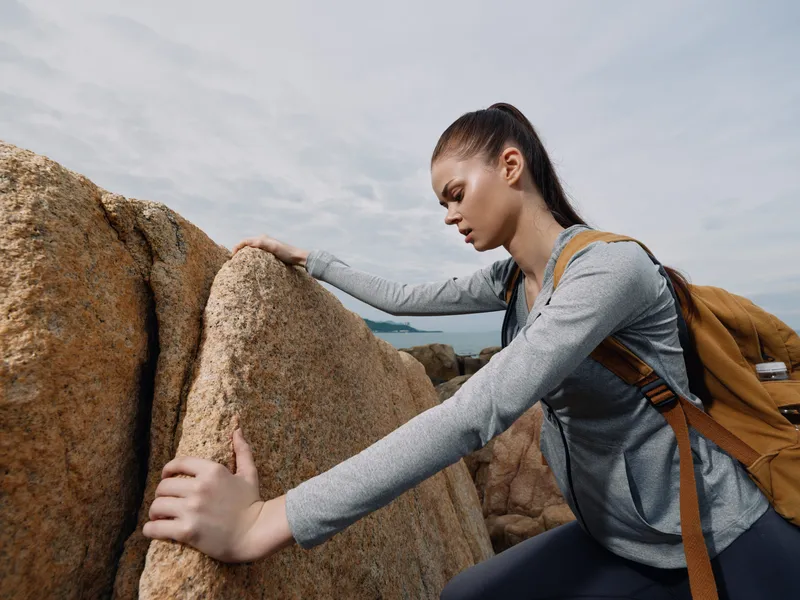 Young woman climbing rocks in athletic wear, showing determination and focus, against a cloudy sky backdrop, perfect for fitness and outdoor adventure themes