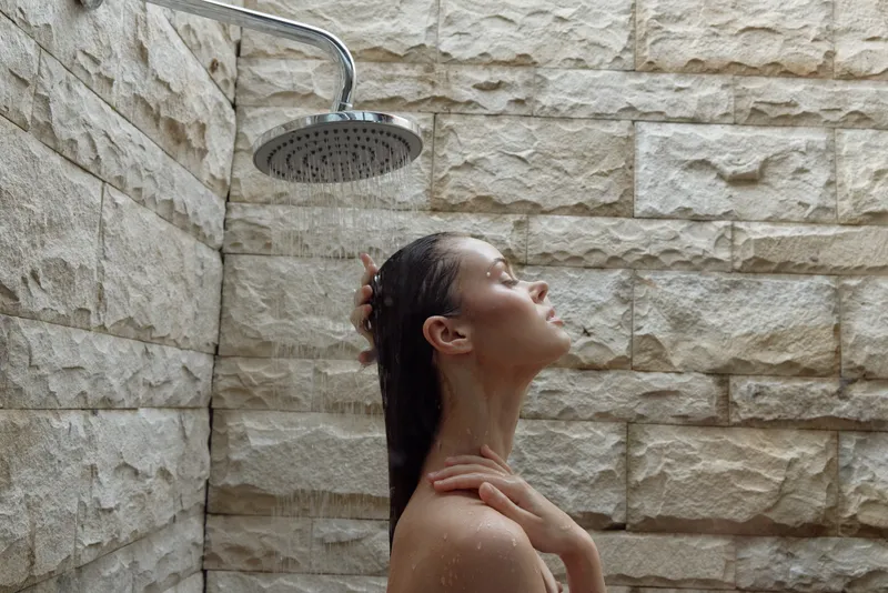 Relaxed woman enjoying shower under rain showerhead in outdoor stone-walled bathroom, natural light enhancing peaceful moment and fresh skin feeling.