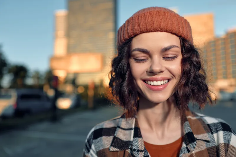 Woman portrait with a warm smile and beanie in urban sunlight, a candid lifestyle frame full of authenticity and golden hour glow, mindful living and emotional storytelling.