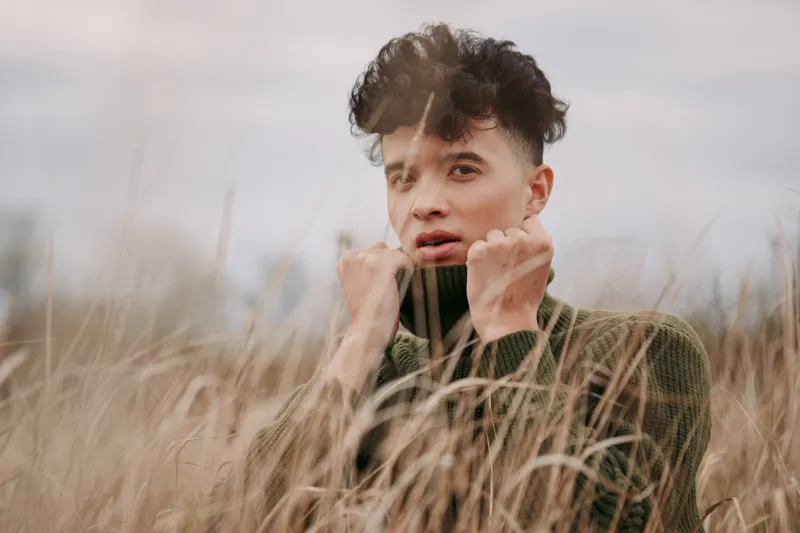 An authentic candid portrait of a young man in tall grass, capturing genuine expression, textured knit, and natural light that emphasizes a believable, real moment.