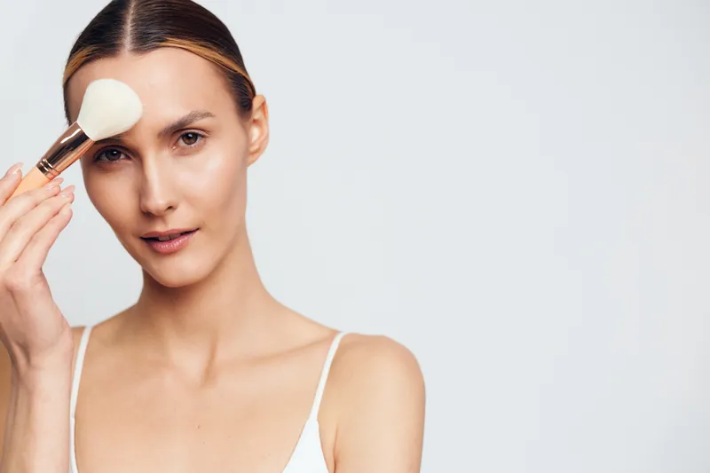 Young woman applying makeup with a brush on her face, clear skin, focused expression, minimal background, beauty routine, natural light, skincare concept.
