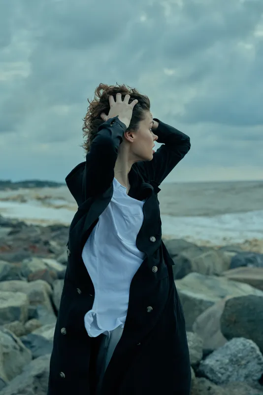 Woman in a long dark coat and white shirt stands on a rocky shore, wind tousling her hair, dramatic sea backdrop, layered clothing, textured fabrics, and candid movement for a bold outdoor fashion